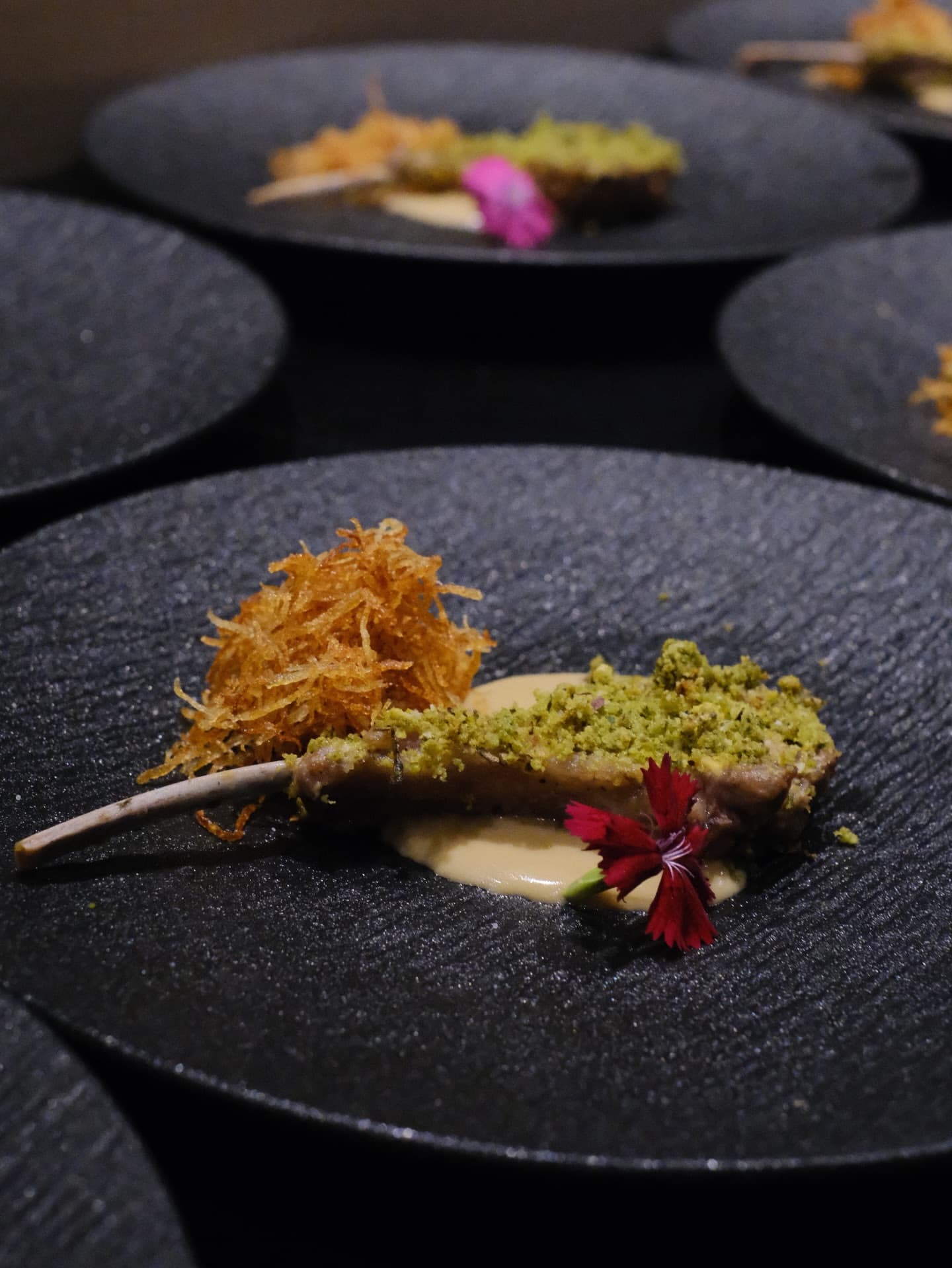 A pistachio-crusted cutlet with a shaved crisp garnish and a flower on a black plate
