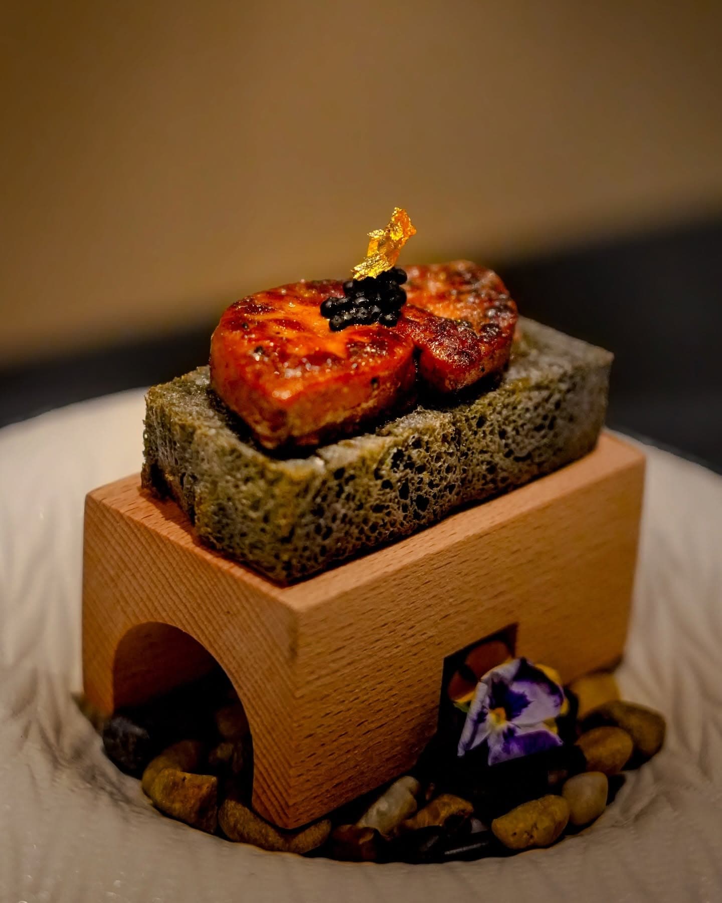 A seared fish course topped with caviar and gold leaf on a green stone block set on a wooden stand